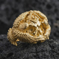 Load image into Gallery viewer, Gold plated Masonic eagle skull ring resting on black volcanic rock, featuring a bold skull centerpiece, raised Square and Compasses symbol, detailed feathered wings, and gothic masculine band design.
