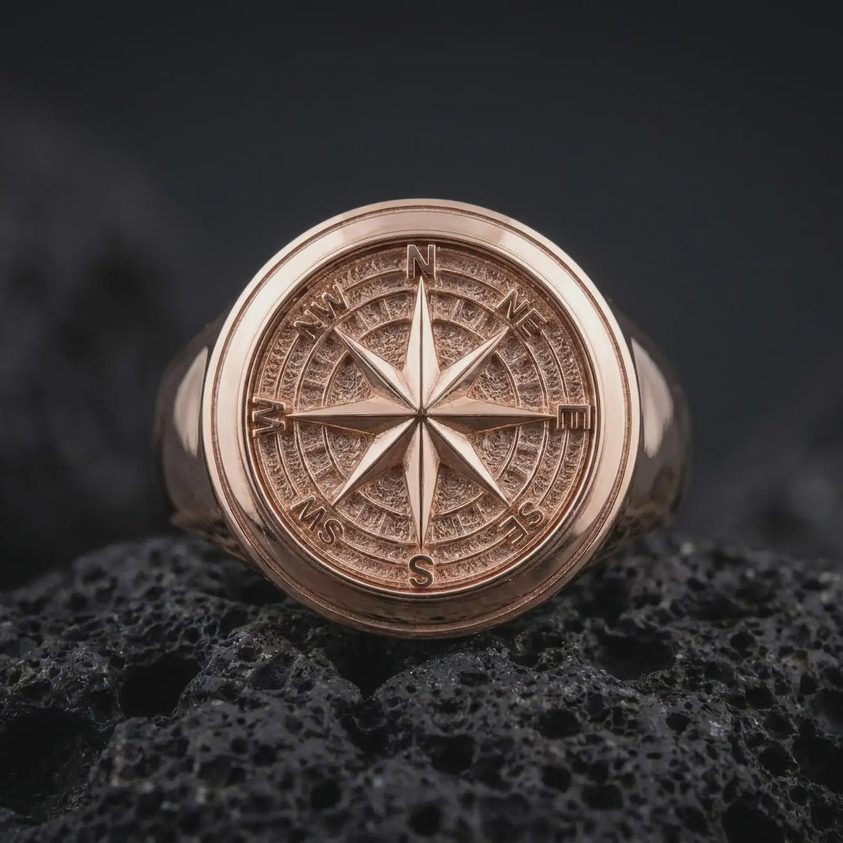 Rose gold plated compass signet ring displayed on textured volcanic stone, showcasing the raised compass rose centerpiece, engraved navigation dial, recessed directional markings, and smooth rounded band.
