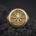Load image into Gallery viewer, Gold plated compass rose ring resting on black volcanic rock, featuring a sculpted eight-point navigation star, detailed directional markers, and a bold circular nautical signet design.
