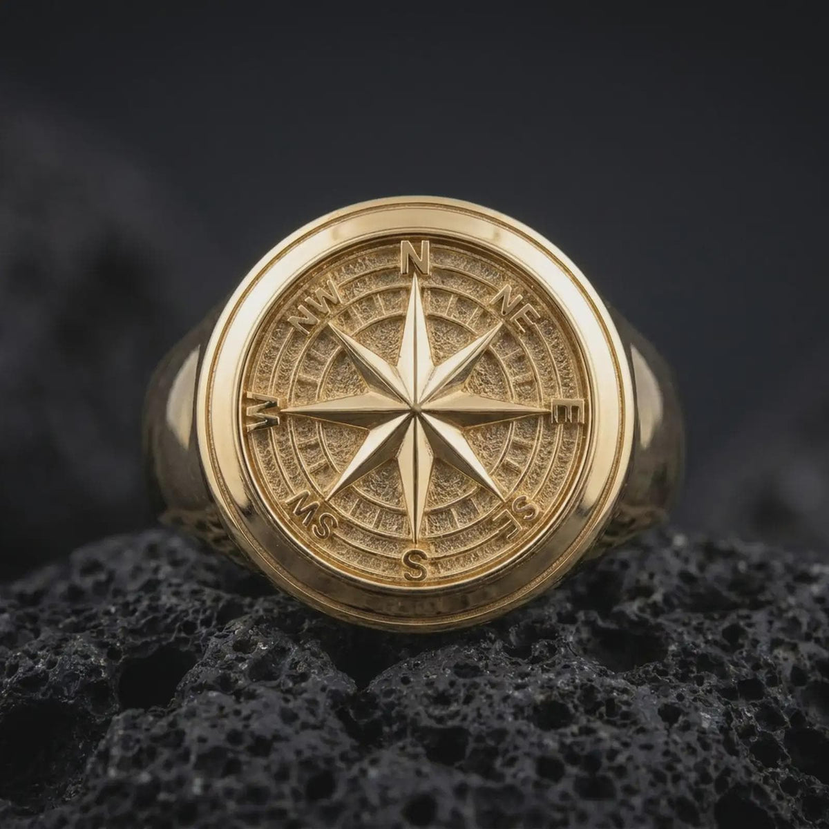 Gold plated compass rose ring resting on black volcanic rock, featuring a sculpted eight-point navigation star, detailed directional markers, and a bold circular nautical signet design.