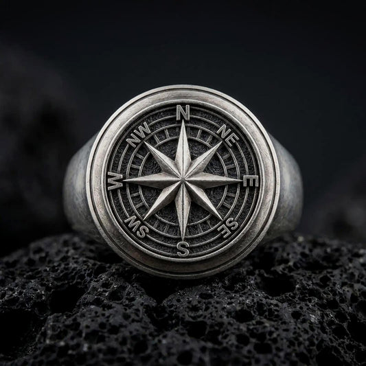 Oxidized sterling silver compass rose signet ring on black volcanic rock, featuring a raised eight-point compass star, engraved directional markings, and a detailed circular navigation dial.