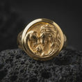 Load image into Gallery viewer, Gold plated lion family signet ring resting on black volcanic rock, featuring a sculpted lion, lioness, and cub embracing in a symbolic family composition with intricate fur details.
