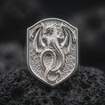 Load image into Gallery viewer, Polished sterling silver winged dragon ring displayed on dark volcanic stone, highlighting the three-dimensional dragon relief, intricate scale patterns, outstretched bat-like wings, and textured medieval shield background.

