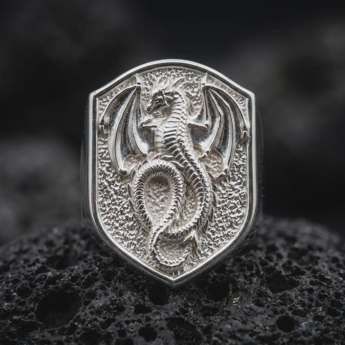 Polished sterling silver winged dragon ring displayed on dark volcanic stone, highlighting the three-dimensional dragon relief, intricate scale patterns, outstretched bat-like wings, and textured medieval shield background.