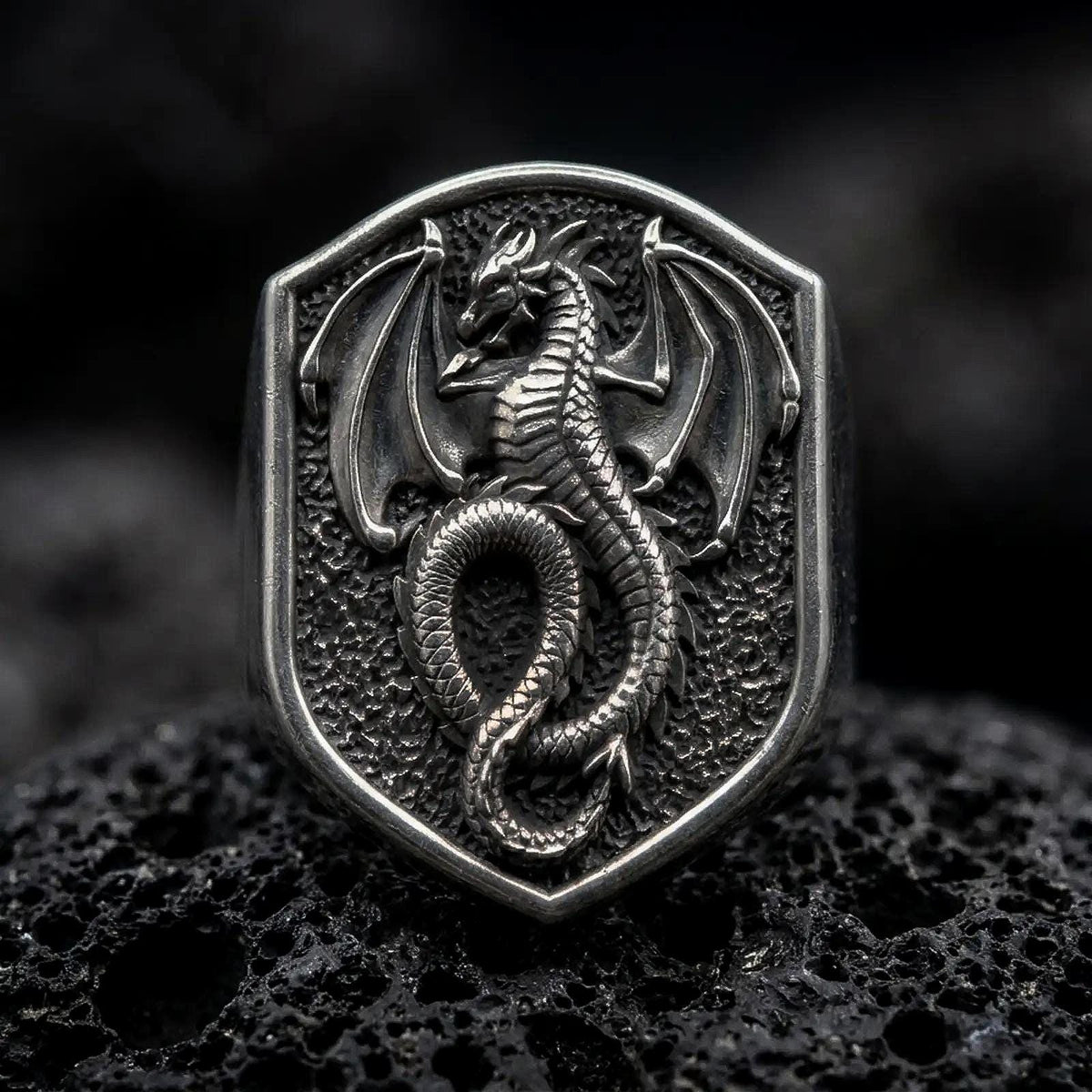 Oxidized sterling silver dragon shield signet ring on black volcanic rock, featuring a raised winged dragon with curved horns, detailed scales, spread wings, and a long coiled tail.