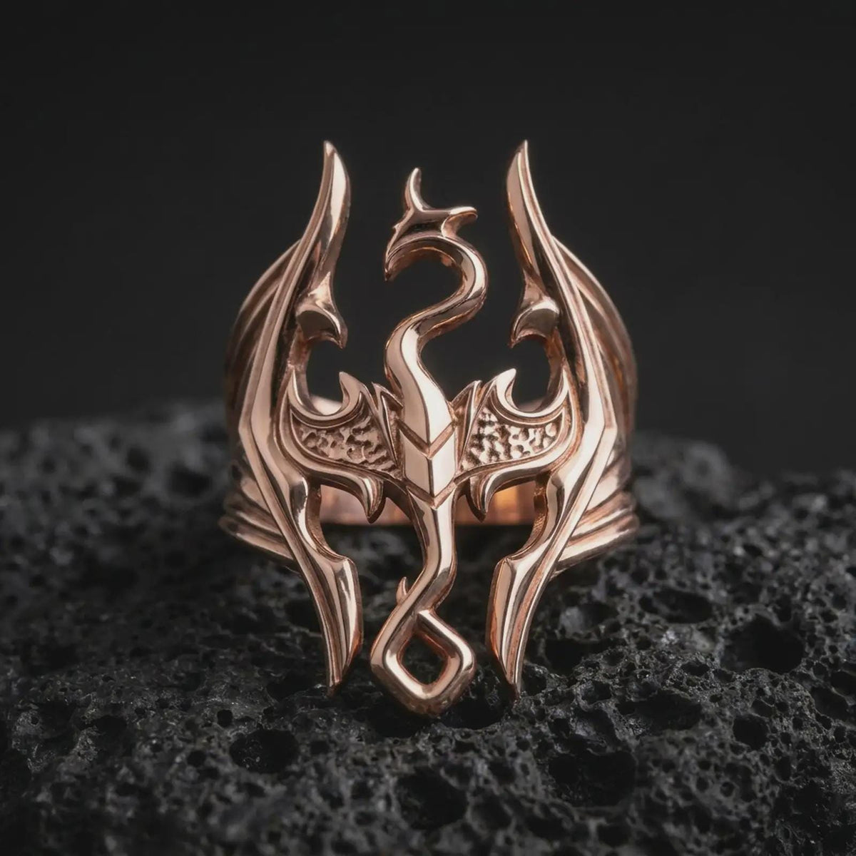 Rose gold plated Elder Scrolls dragon ring displayed on textured volcanic stone, showcasing the raised dragon silhouette, sweeping pointed wings, detailed central body, and bold Dovahkiin-inspired design.