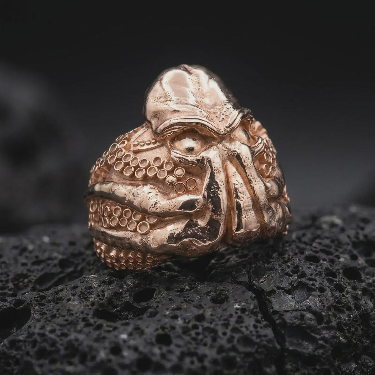 Rose gold plated Lovecraftian monster ring displayed on textured volcanic stone, showcasing the sculpted Cthulhu face, deep-set eyes, thick octopus tentacles, and bold cosmic horror-inspired statement design.
