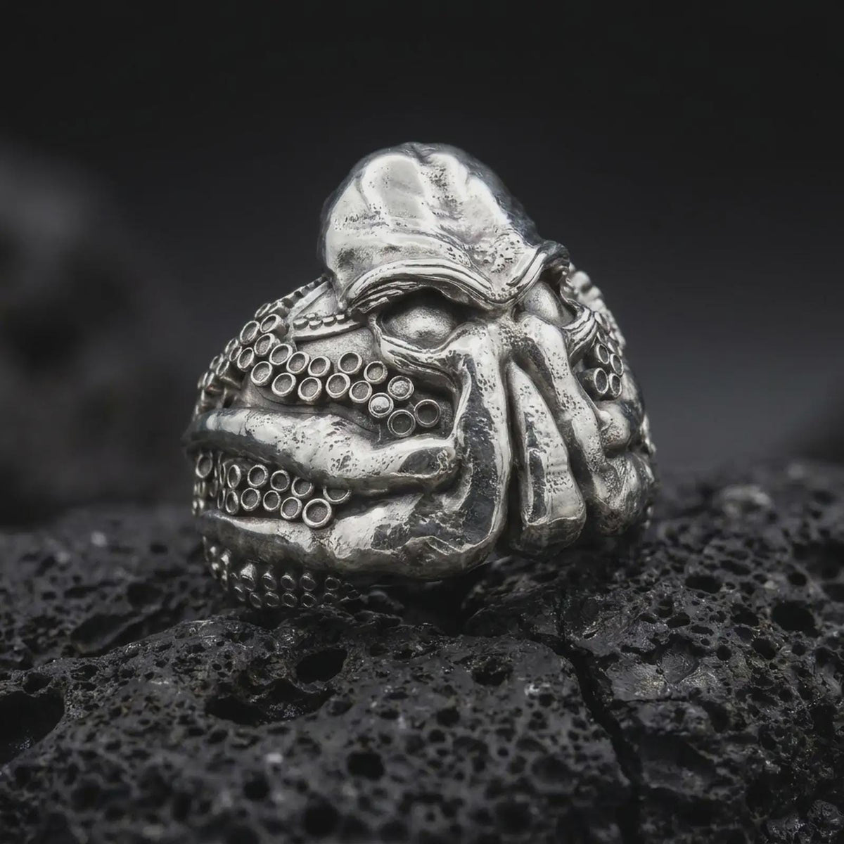Polished sterling silver Lovecraft Cthulhu ring displayed on dark volcanic stone, highlighting the massive sculpted forehead, sinister eye sockets, curling tentacles, and intricate circular sucker patterns wrapping around the band.