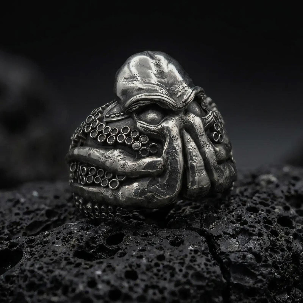 Oxidized sterling silver Cthulhu ring on black volcanic rock, featuring a monstrous Lovecraftian face, deeply recessed eyes, thick facial tentacles, and detailed octopus suction cups.