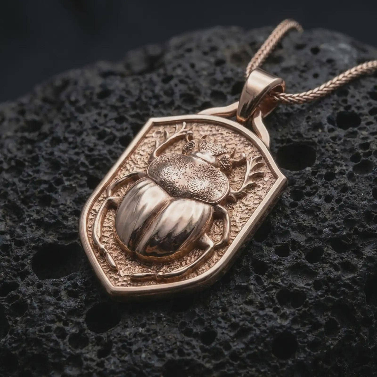 Rose gold plated beetle shield necklace displayed on textured volcanic stone, showcasing the dimensional stag beetle sculpture, intricate body textures, branching mandibles, and raised shield border.