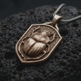 Load image into Gallery viewer, Bronze stag beetle shield pendant on black volcanic rock, featuring a raised beetle with antler-like mandibles, detailed wing covers, sculpted legs, and a textured shield background.
