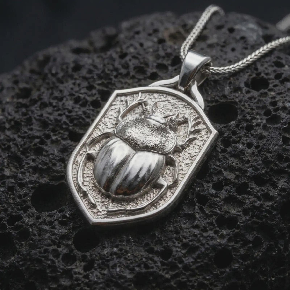 Polished sterling silver stag beetle necklace displayed on dark volcanic stone, highlighting the three-dimensional beetle body, curved mandibles, segmented wing covers, and shield-shaped medallion frame.