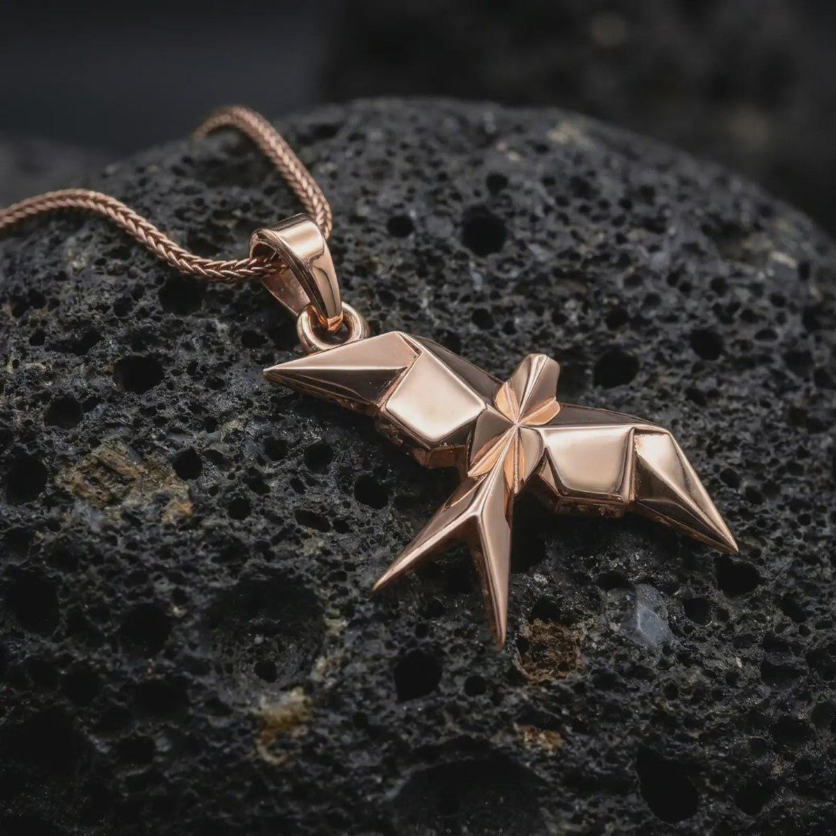 Rose gold plated geometric swallow necklace displayed on textured volcanic stone, showcasing the folded-paper inspired form, angular wings, and minimalist sculptural design.