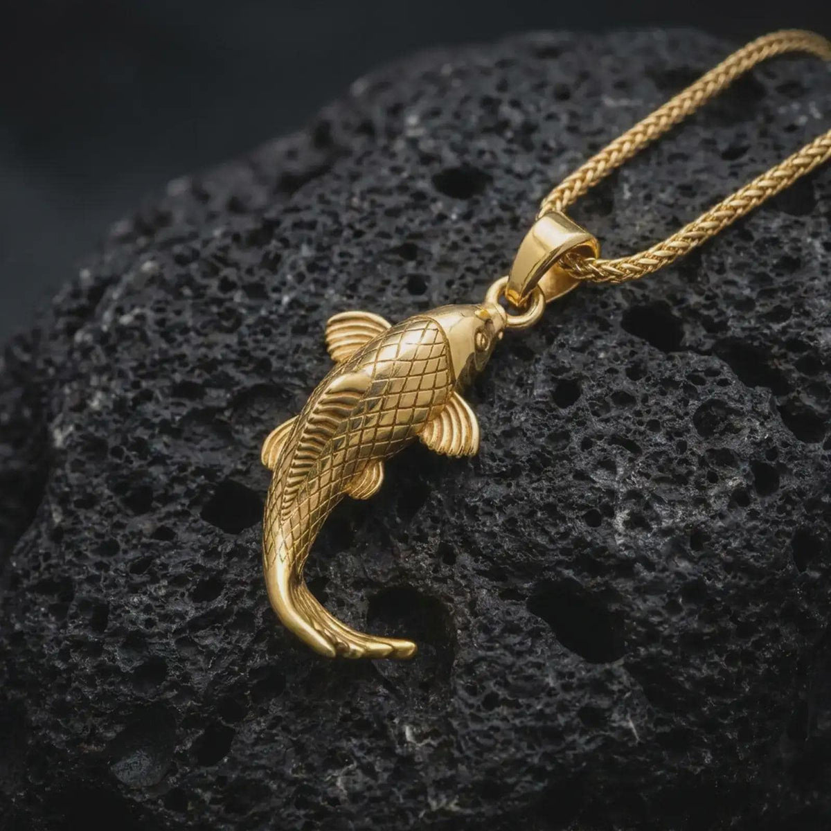 Gold plated Japanese koi fish pendant resting on black volcanic rock, featuring detailed scales, curved tail, sculpted fins, and an elegant lucky carp design.