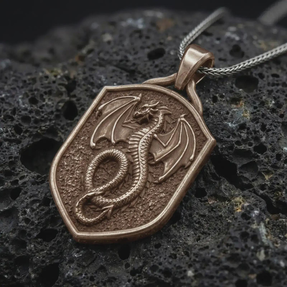 Bronze coiled dragon shield pendant on black volcanic rock, featuring a winged dragon, detailed scales, curled serpentine body, and textured medieval shield.