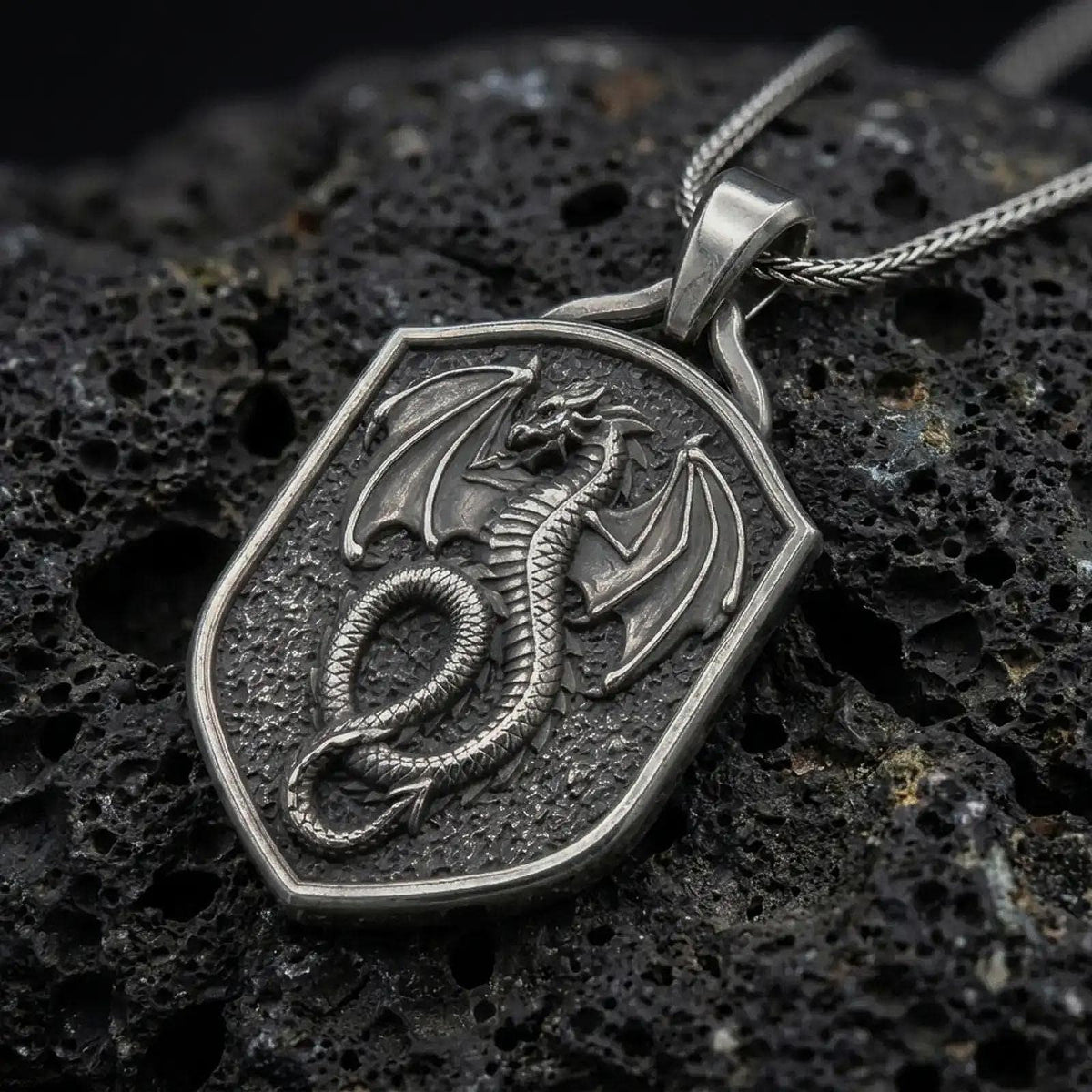 Oxidized sterling silver coiled dragon shield pendant on black volcanic rock, featuring a winged dragon, detailed scales, curled serpentine body, and textured medieval shield.
