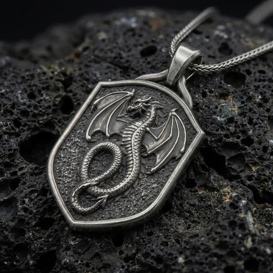 Oxidized sterling silver coiled dragon shield pendant on black volcanic rock, featuring a winged dragon, detailed scales, curled serpentine body, and textured medieval shield.