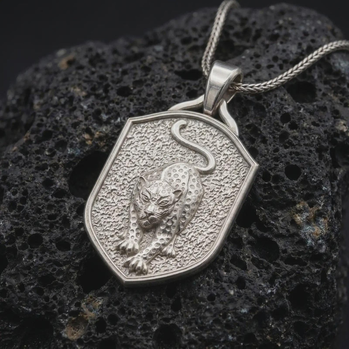 Polished sterling silver leopard pendant displayed on dark volcanic rock, highlighting the three-dimensional wild cat sculpture, detailed spotted coat, muscular shoulders, focused eyes, and raised shield-shaped silver frame.