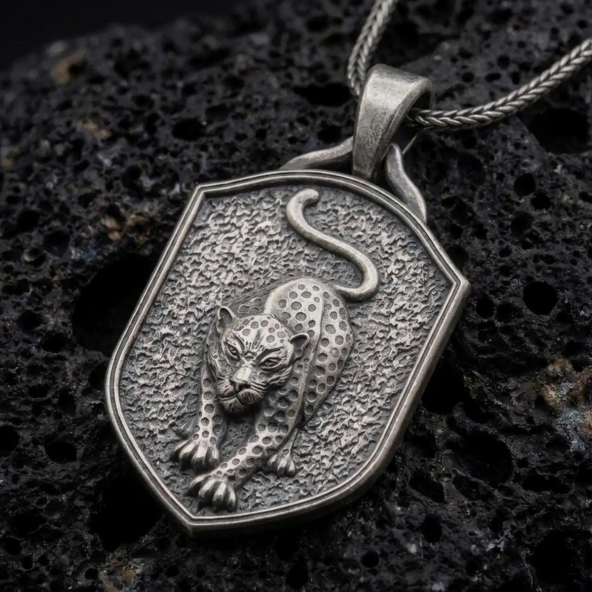 Oxidized sterling silver leopard pendant necklace on black volcanic stone, featuring a prowling spotted leopard with extended front paws, curved tail, textured shield-shaped background, and braided silver chain.