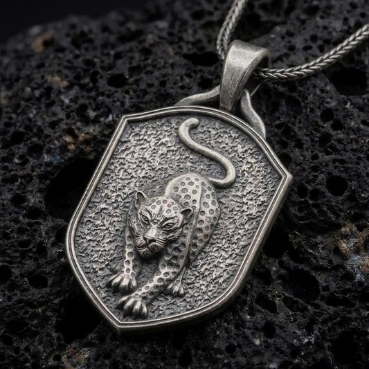 Oxidized sterling silver leopard pendant necklace on black volcanic stone, featuring a prowling spotted leopard with extended front paws, curved tail, textured shield-shaped background, and braided silver chain.