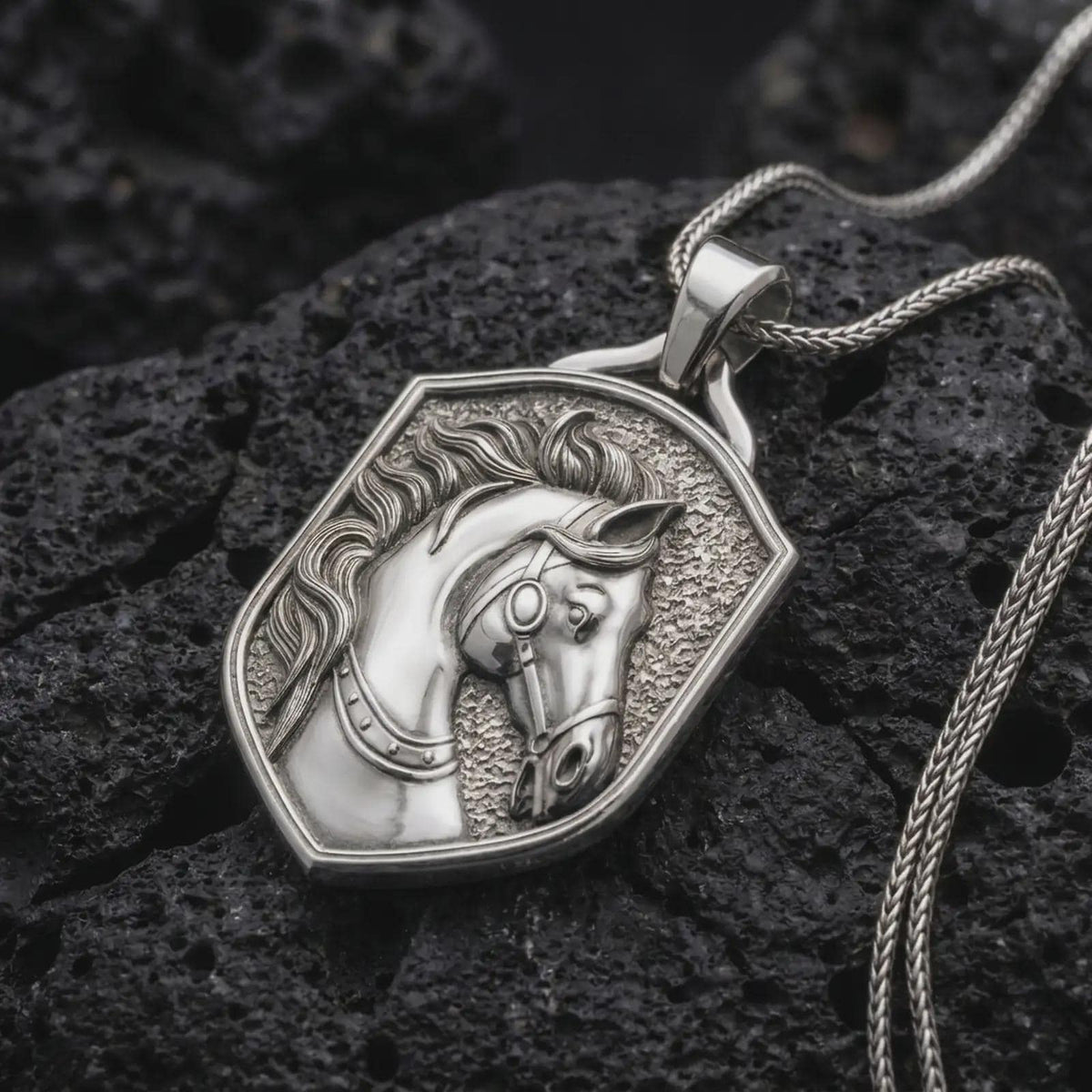 Polished sterling silver equestrian horse pendant displayed on dark volcanic rock, highlighting the three-dimensional horse portrait, windswept mane, decorative bridle straps, circular cheekpiece, and detailed facial features.