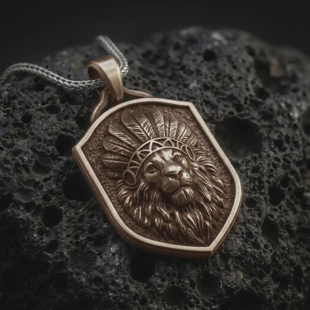 Bronze lion chief pendant necklace on black volcanic stone, featuring a majestic lion head wearing a detailed feather headdress, flowing mane, textured shield-shaped background, and braided silver chain.