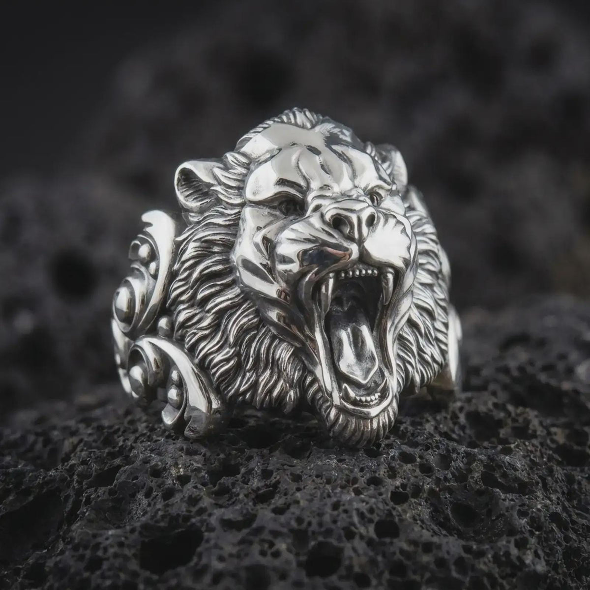 Polished silver tiger head ring on dark lava stone, showcasing a fierce roaring expression, deep-set eyes, pronounced nose, textured mane-like fur, and statement ring silhouette.
