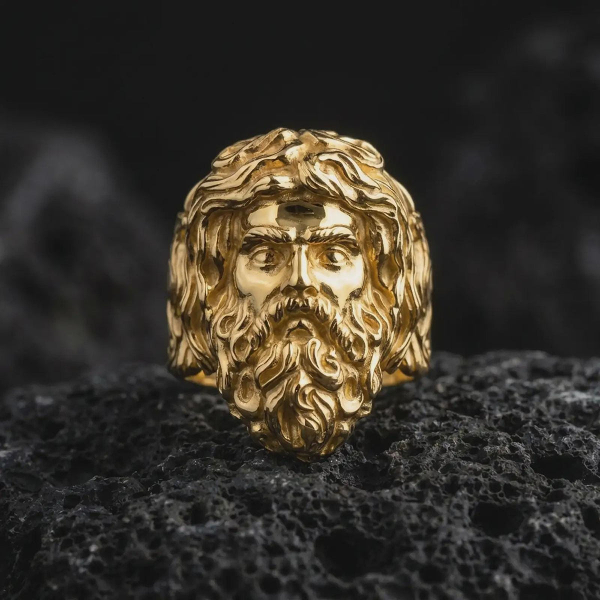 Gold plated sterling silver Poseidon ring displayed on dark volcanic rock, featuring a raised ancient Greek sea god face with an elaborate wavy beard, flowing hair, powerful facial expression, and warm gold finish.
