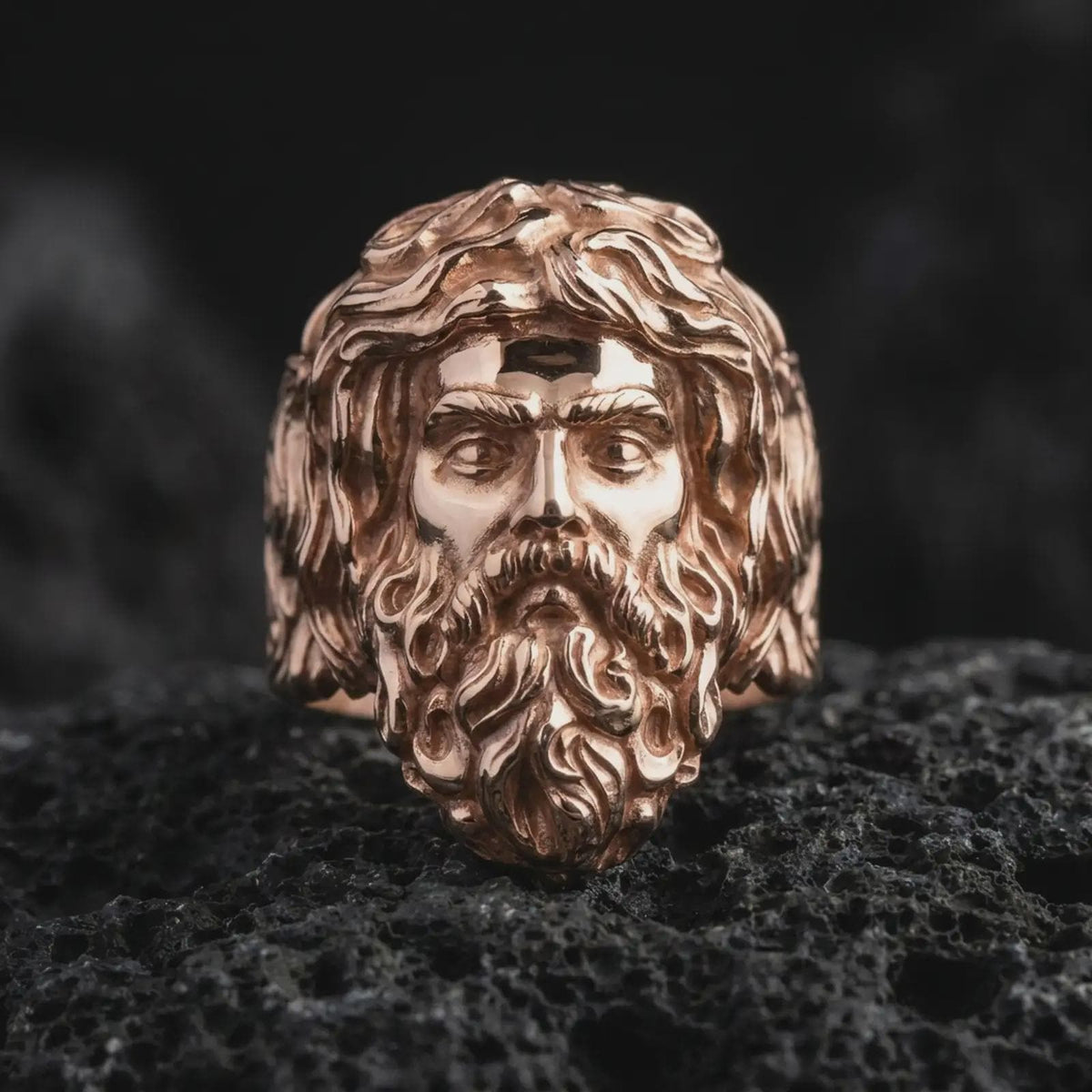 Rose gold plated sterling silver Poseidon ring resting on black volcanic stone, showcasing the dimensional Greek mythology god portrait, detailed facial anatomy, flowing beard, sculpted hair, and elegant rose gold finish.