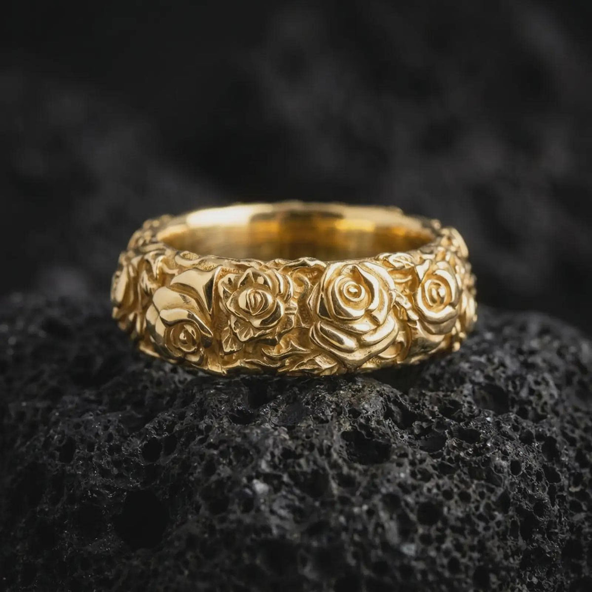 Gold plated sterling silver rose flower band ring displayed on dark volcanic rock, featuring detailed three-dimensional roses, overlapping petals, botanical leaf engravings, and a warm gold plated finish.