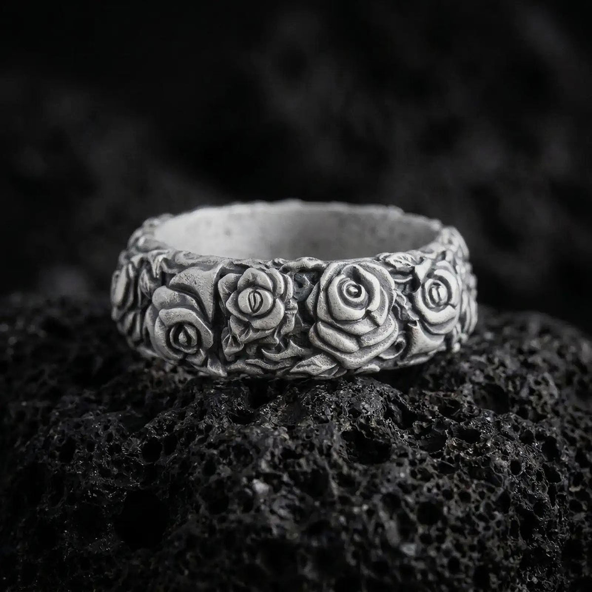 Oxidized 925 sterling silver rose flower band ring displayed on black volcanic rock, featuring three-dimensional rose blossoms, layered petals, sculpted leaves, dark recessed floral details, and a wide ornamental band.