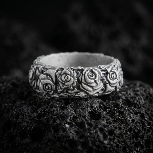 Oxidized 925 sterling silver rose flower band ring displayed on black volcanic rock, featuring three-dimensional rose blossoms, layered petals, sculpted leaves, dark recessed floral details, and a wide ornamental band.