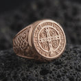 Load image into Gallery viewer, Rose gold Saint Benedict ring resting on black volcanic stone, highlighting the circular Catholic protection medal, traditional cross and Latin inscriptions, decorative Celtic scrollwork, and warm rose gold plated finish.
