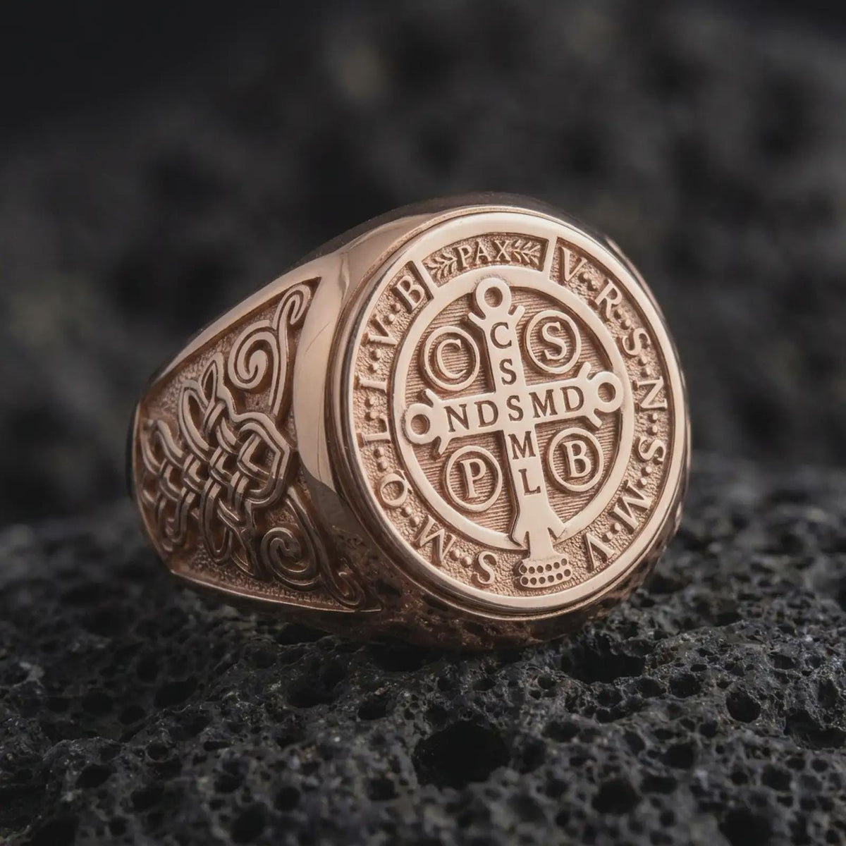 Rose gold Saint Benedict ring resting on black volcanic stone, highlighting the circular Catholic protection medal, traditional cross and Latin inscriptions, decorative Celtic scrollwork, and warm rose gold plated finish.