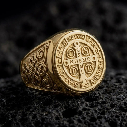 Gold Saint Benedict Medal ring displayed on dark volcanic rock, featuring the detailed Benedictine Cross, engraved Latin initials, PAX inscription, ornate Celtic knotwork, and a bright polished gold finish without blackened recesses.