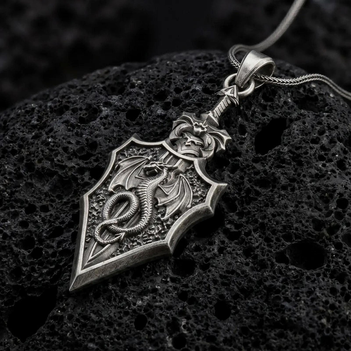 Oxidized 925 sterling silver dragon sword pendant displayed on textured black volcanic rock, featuring a winged dragon coiled around a medieval-style sword blade, intricate scales, decorative hilt, angular edges, and dark oxidized silver finish.