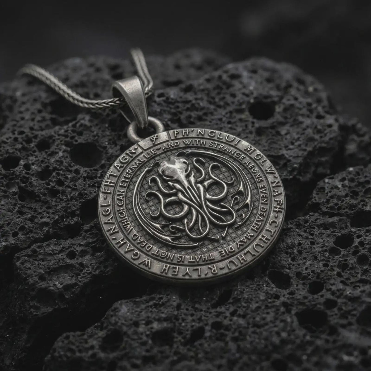 Oxidized sterling silver Cthulhu pendant on black volcanic rock, featuring a raised cephalopod sigil in the center, engraved outer ring lettering, and a dark Lovecraftian medallion design.