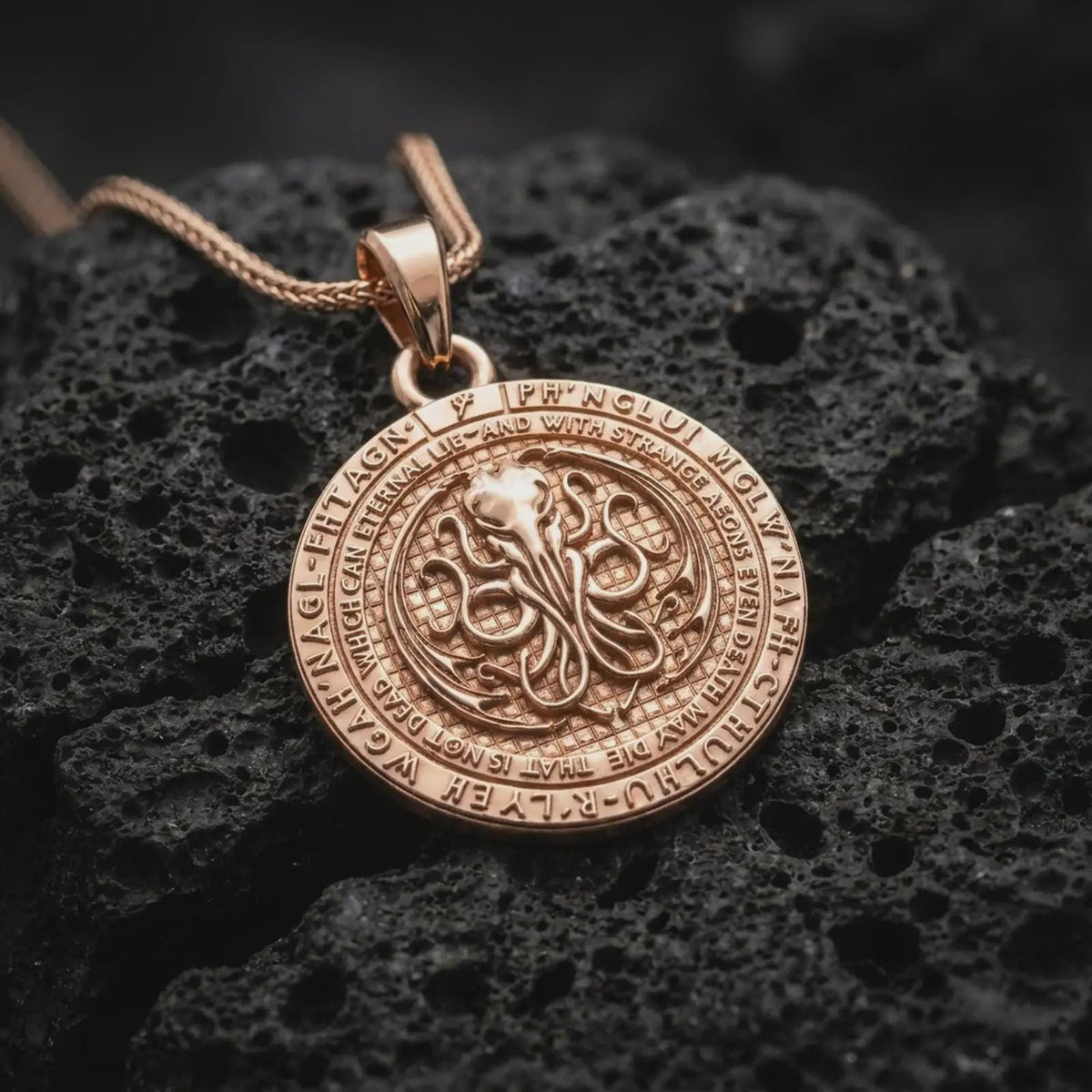 Rose gold plated Lovecraftian amulet displayed on dark volcanic stone, showcasing the raised cephalopod emblem, engraved fantasy lettering, and gothic statement pendant style.