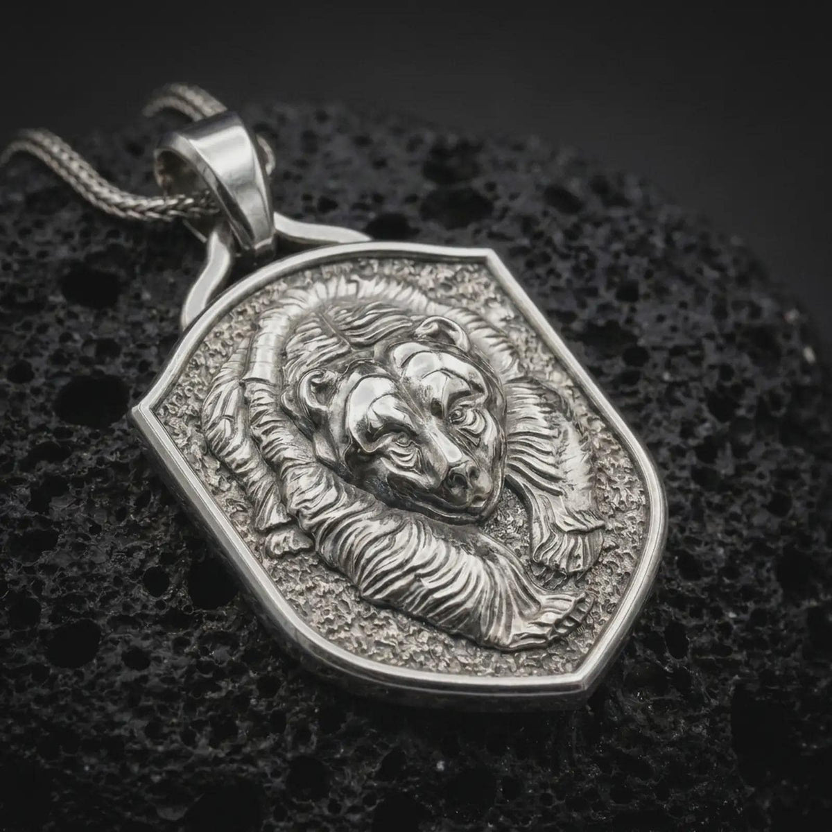 Polished sterling silver bear necklace displayed on dark volcanic stone, highlighting the detailed grizzly face, flowing fur texture, powerful expression, and shield-shaped pendant frame.