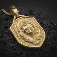 Load image into Gallery viewer, Gold plated grizzly bear pendant resting on black volcanic rock, featuring a raised bear portrait, detailed fur, rugged textured field, and bold heraldic shield silhouette.
