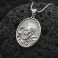 Load image into Gallery viewer, Polished sterling silver Saint Padre Pio medal displayed on dark volcanic stone, highlighting the sculpted saint portrait, raised hand, circular halo, textured oval background, and cross-decorated bail.
