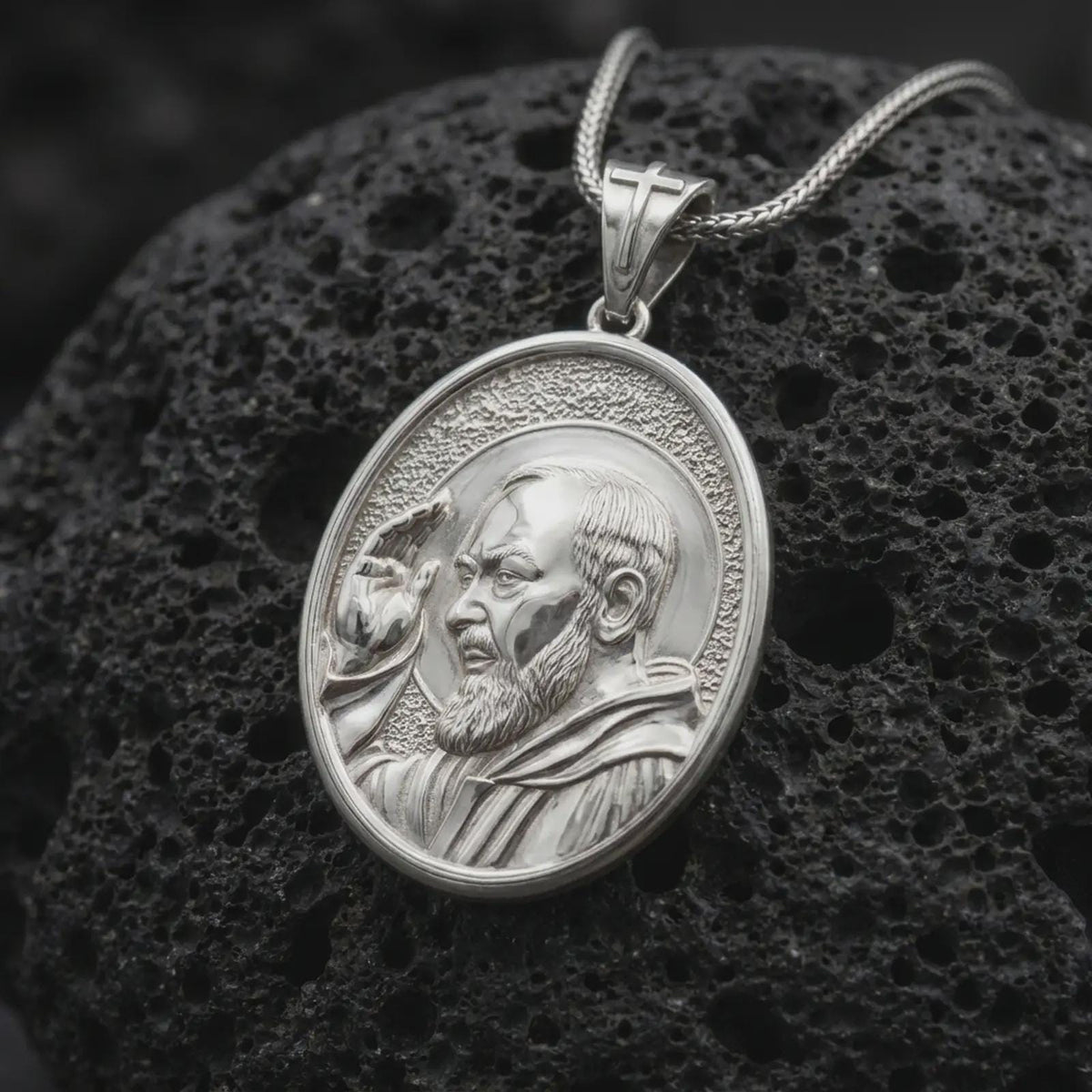 Polished sterling silver Saint Padre Pio medal displayed on dark volcanic stone, highlighting the sculpted saint portrait, raised hand, circular halo, textured oval background, and cross-decorated bail.