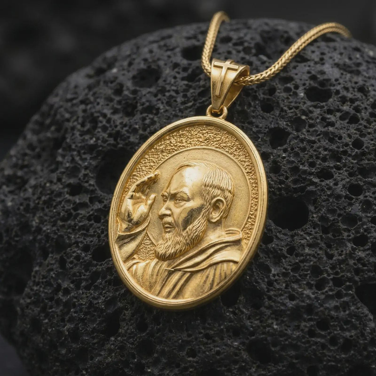 Gold plated Padre Pio necklace resting on black volcanic rock, featuring the detailed Catholic saint portrait, traditional Capuchin robe, blessing gesture, and oval religious medallion design.