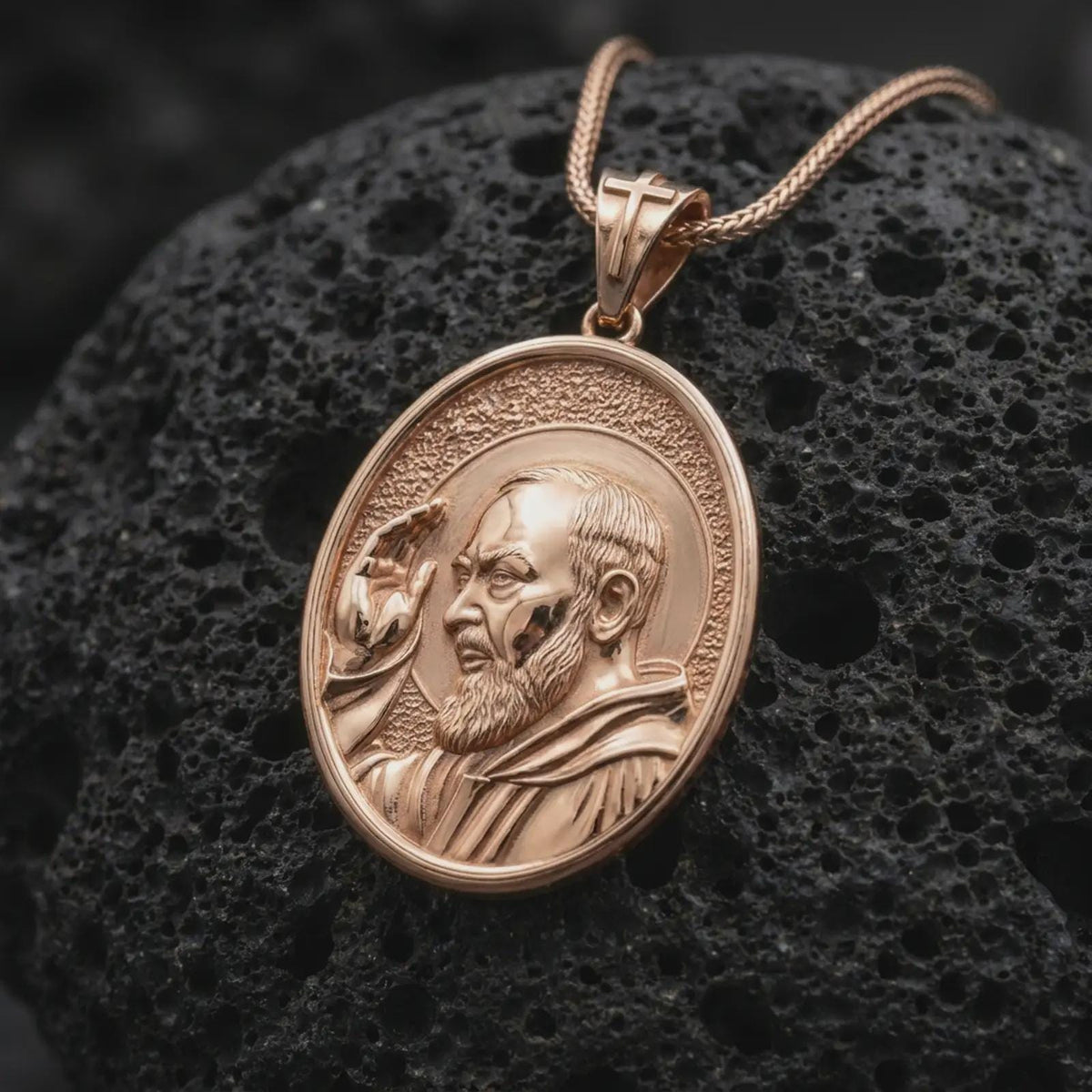 Rose gold plated Saint Pio pendant displayed on textured volcanic stone, showcasing the realistic facial relief, detailed beard, raised blessing hand, halo, and meaningful Catholic devotional jewelry design.