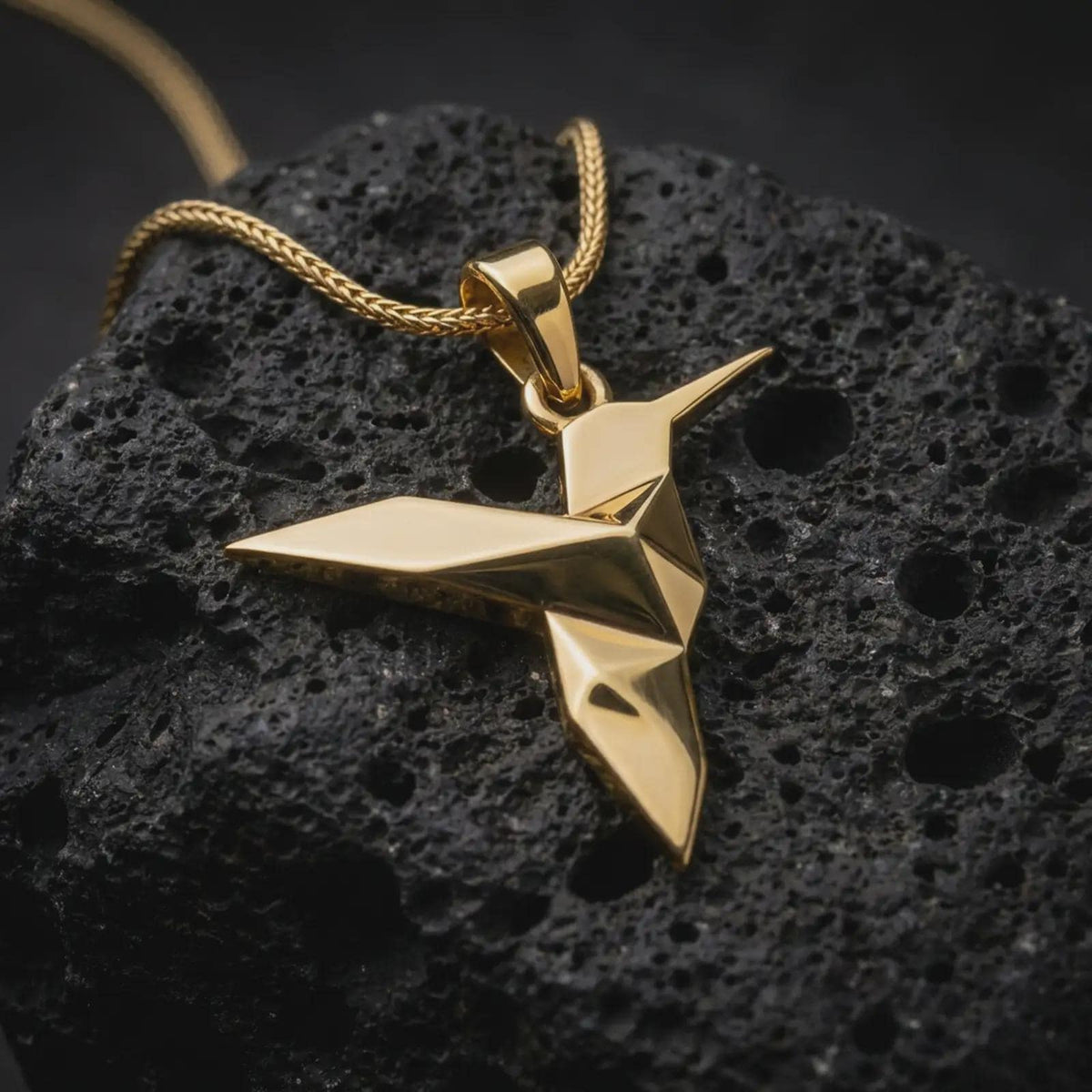 Gold plated origami hummingbird pendant resting on black volcanic rock, showcasing the sleek geometric bird shape, pointed wings, and contemporary nature inspired jewelry design.