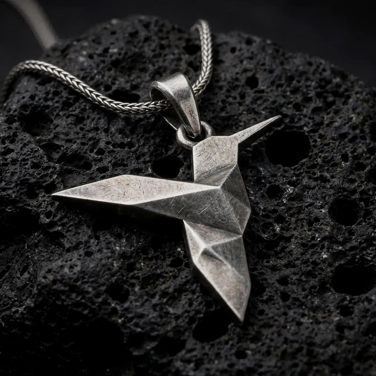 Oxidized sterling silver origami hummingbird pendant on black volcanic rock, featuring a geometric folded bird design with a long pointed beak and angular wings.