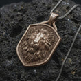 Load image into Gallery viewer, Bronze regal lion shield pendant on black volcanic rock, featuring a majestic lion portrait, calm expression, flowing mane, and textured heraldic shield background.
