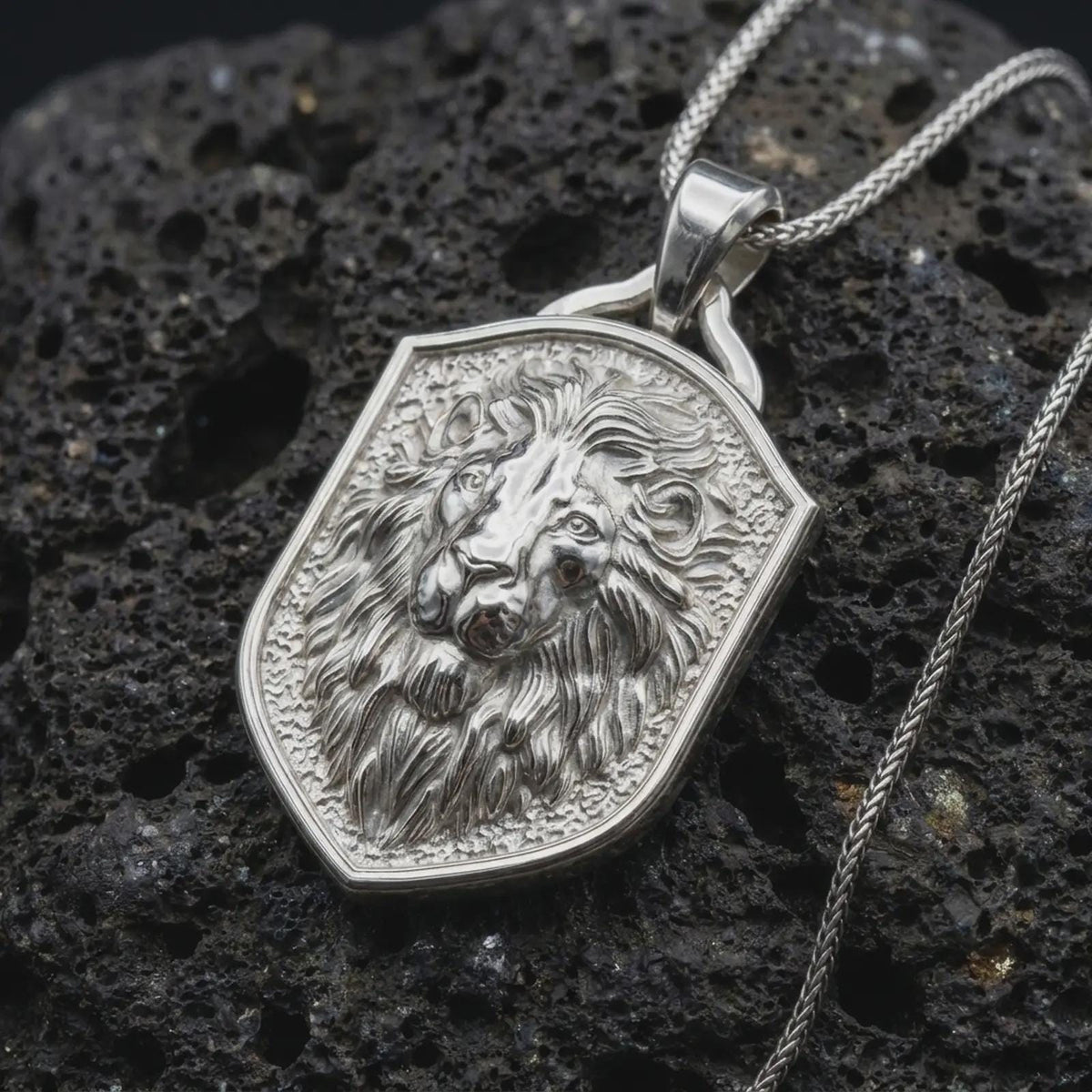 Polished sterling silver lion head necklace displayed on dark volcanic stone, highlighting the sculpted lion face, expressive eyes, detailed flowing mane, and shield-shaped medallion frame.
