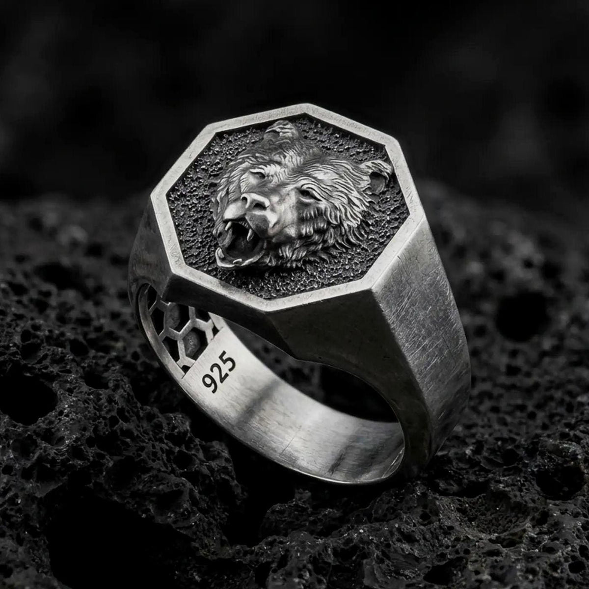 Oxidized 925 sterling silver roaring bear signet ring displayed on textured black volcanic rock, featuring an open-mouthed bear head, sharp fangs, detailed fur, hexagonal frame, and dark oxidized silver finish.