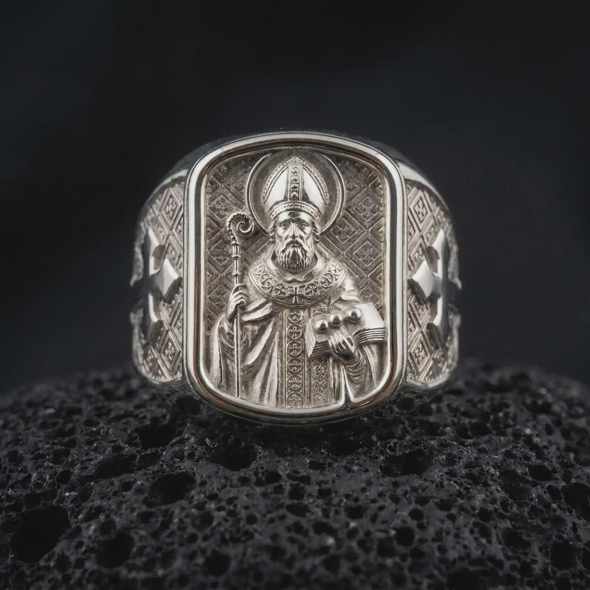 Polished 925 sterling silver St Nicholas Catholic ring resting on black volcanic stone, showcasing a sculpted bishop portrait with a circular halo, decorated mitre, curved pastoral staff, three balls on a book, and bright polished silver finish.